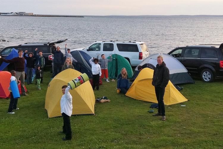 Informal photo of choir members standing with camping tents and vans at seaside during tour 