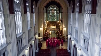 Photo taken from high up in Cathedral clerestory windows, looking down the nave towards high altar and stained glass window, with the choir in red robes in the crossing.